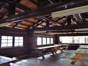 Inside a large wooden picnic shelter. The ceiling is held up by large wooden beams. The walls are log cabin construction with many windows. The floor is cement. There are several picnic tables. 
